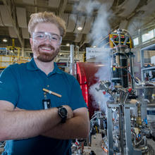 Young man with wild blonde hair and reddish beard smiling beside an AC susceptometer that he helped build