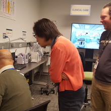 One person sits in front of a wireless infusion pump. Two people stand and look over his shoulder. Other pumps are in the background.