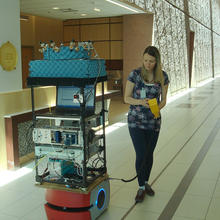 Woman in large indoor hallway standing next to tall stack of electronic equipment.