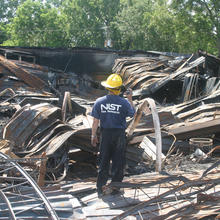 Man in NIST fire research t-shirt and hard hat standing among wreckage
