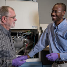 Two men (Bob Vocke and Savelas Rabb )stand in a lab setting, smiling and discussion something in front of their mass spectrometer, a large instrument used in chemistry.