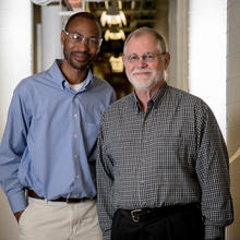 Two men (Bob Vocke and Savelas Rabb) stand in a hallway outside of a lab. They are both smiling