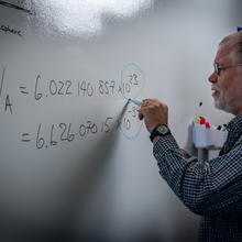 Chemist Bob Vocke stands in front of a white board writing out several complicated equations related to the mole redefinition.