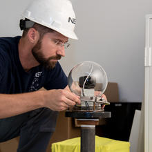 man with a beard, safety glasses, hard hat and blue shirt places a glass bubble over the top of a camera mounted on a steel pole inside the test room