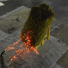 A rectangular-shaped model of a thatched roof is attacked by embers emitted from the pipe-shaped mouth of the NIST Dragon device.