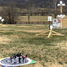 An aerial drone sits on a launch pad before lifting off to start the NIST performance test course. Three sections of the course, poles with white bucket-shaped targets attached, are seen in the background.