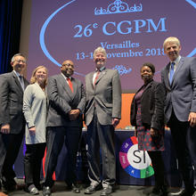 6 people standing on a stage in front of a screen that reads: 26th CGPM