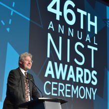 A man stands at a podium in front of a large audience, and behind him a screen is lit up with the words 46th Annual NIST Awards Ceremony."