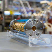 cylindrical rod of metal-banded disks capped by aluminum donuts resting on a metal table. A band of blue circuitry runs along the top.  