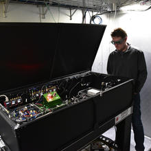 Man in goggles standing next to and looking into a large box with open lid showing what appears to be electrical equipment and wiring inside