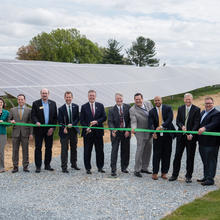 Eleven people stand in front of the NIST solar array getting ready to cut an orange ribbon with large scissors.