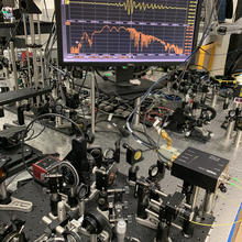 Table covered with lenses and other optical equipment, and a flat screen mounted high in the background showing a yellow squiggly line across the top and a spiky orange line below.