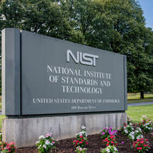 gray entrance sign that says NIST National Institute of Standards and Technology, United States Department of Commerce, 100 Bureau Drive. Surrounded by green grass, flowers, trees.