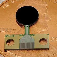 A small flat device with a round black part and a copper-colored rectangular part lies on top of a penny.