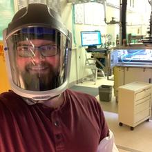 A man wearing a safety visor poses in front of lab equipment. 