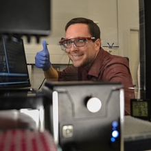 A man in safety goggles gives a thumbs-up while sitting at a computer.