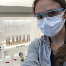 A woman wearing safety goggles and a mask poses in front of a cabinet holding vials.