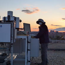 Man standing next to equipment on top of building during a sunset.  