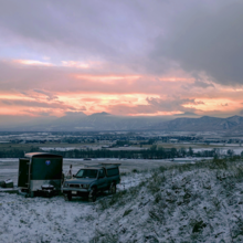 A small pickup truck and trailer stand on a grassy slope with snow and mountains in the background.