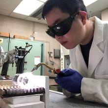 A man (Zhi Liang) wearing dark safety goggles leans over a table, working with scientific equipment.