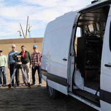 Four people stand around an instrument on a tripod next to the open door of a white van.