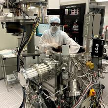 Nathan Tomlin wears protective clothes and safety goggles while reaching into a complex-looking scientific device. 