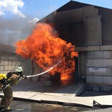 Firefighters spray water into a burning concrete building with a sign saying "NIST Fire Research."