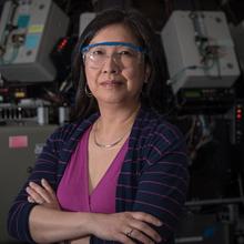 Joannie Chin stands with arms crossed, wearing safety goggles, in front of scientific equipment. 
