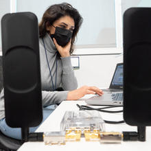A woman wearing a face mask works at a laptop with communications equipment on the desk. 
