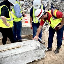 Four workers in hard hats look down at a damaged concrete pillar. 