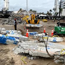 Pieces of concrete marked with blue spray paint lay on the ground amid pieces of construction equipment. 