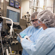 Two people in coveralls and hair covers check the readouts on laboratory equipment.