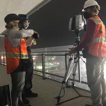 Three people in orange vests stand on a darkened balcony near a tripod.