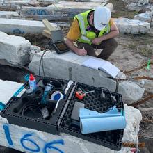 A man in a hard hat crouches next to a concrete column, taking measurements.