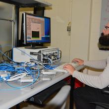 Man with beard and glasses in light grey sweater sitting on the right looking at computer screen on the left, with mass of blue wires in the foreground.