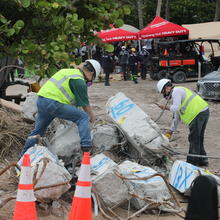 Two people in hard hats and other safety gear bend over broken concrete columns marked with blue spray paint, with tents and construction equipment in the background. 