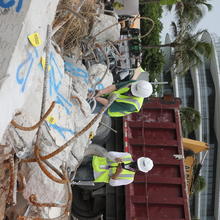 Two people in hard hats and other safety gear examine broken concrete columns with a dumpster and a condominium building in the background. 