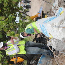 A man in a hard hat and other safety gear takes a picture of a broken concrete column with his phone.