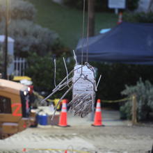 A piece of broken concrete column is lifted by a crane with construction equipment in the background.