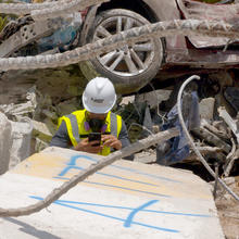 A person in a hard hat surrounded by debris including a crushed car takes a picture of a concrete block with a phone.