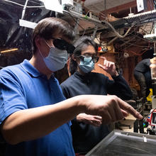 Jun Ye in mask on left pointing at a device. Man in mask stands beside him.