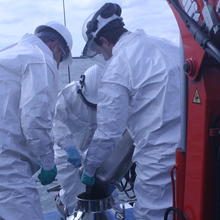 Three researchers on a boat wearing white overalls gather around to help pour material into a metal container.