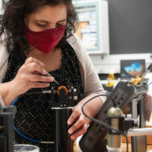 A woman wearing a face covering leans over a piece of scientific equipment, holding tweezers. 