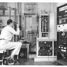Historical photo shows a man dressed in white sitting on a stool working with a wall of scientific equipment.
