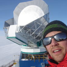 A man in winter gear stands in front of a large dish with a background of snow and blue sky. 