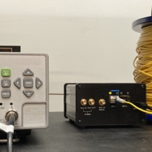 A small rectangular device with a display screen and buttons sits next to a spool of yellow fiber. 