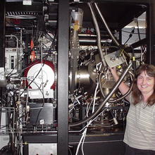 A woman stands smiling in front of a large device with many metal parts and wires.