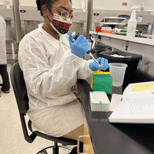 A woman (Diane Nelson) in a mask and lab coat works with plastic test tubes at a lab bench.