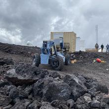 A blue construction vehicle sits near a pile of rubble with three people in the background.