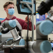 A man wearing a face mask touches a computer screen in a lab with robotic arms in the foreground.